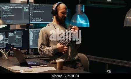 Programmer being happy and playing air guitar with keyboard instrument, enjoying music or song and having fun. Young cheerful adult acting silly fooling around late at night in office. Stock Photo