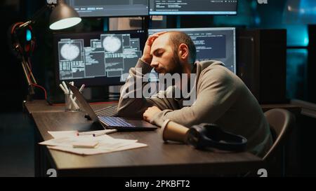 App IT engineer sleeping on desk and being drained, trying to work with artificial intelligence at night. Tired sleepy man yawning and falling asleep in office, using multiple monitors. Stock Photo