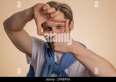 Man photographer gesturing picture frame with hands, looks through fingers and focusing on interesting moment, imitating zoom and cropping nice image. Young guy isolated on beige studio background Stock Photo