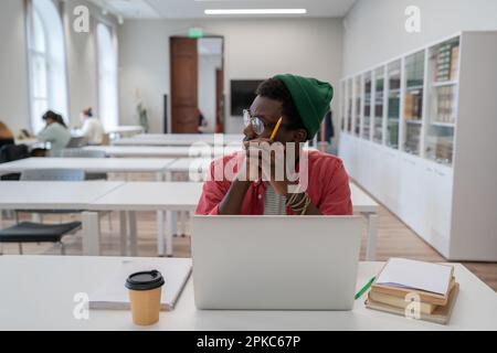 Thoughtful african american nerd man student studying in library using laptop. Stock Photo