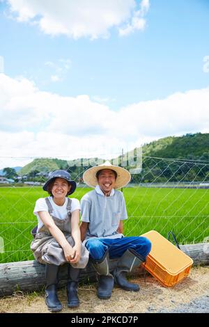 Smiling man and woman sitting on a log in front of a paddy field Stock Photo