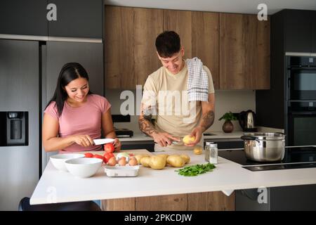 Two young latin people peeling potatoes and cutting tomatoes at kitchen. Stock Photo