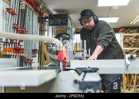 Focused professional male craftsman saws wood on a circular saw in protective eyewear and headphones. Woodworking studio interior. High quality photo Stock Photo