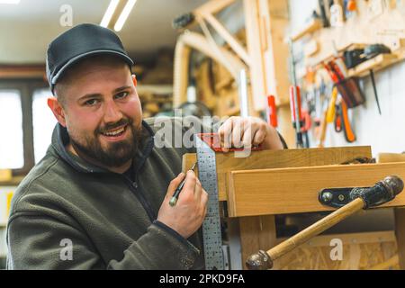 Smiling positive male carpenter using measuring bracket and pencil to create another wooden art piece. Carpentry studio. High quality photo Stock Photo