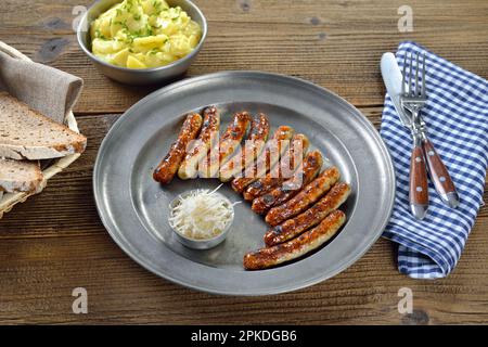 Ten fried Nuremberg sausages with fresh horseradish served on a pewter plate with a bowl of potato salad and farmers bread Stock Photo