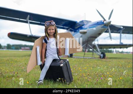 A little girl in a pilot's costume sits on a retro suitcase at the airport waiting for the departure of the flight. A child in a hat and glasses is Stock Photo