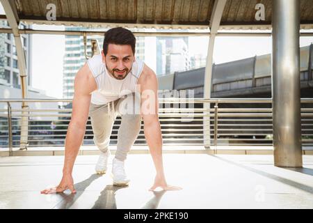 Asian sports running man working out and stretch warm up leg muscle before outdoor running exercise along urban city after business work. Outdoor fitn Stock Photo