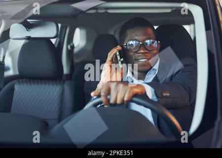 Front view. Young african american businessman in black suit is in the automobile. Stock Photo