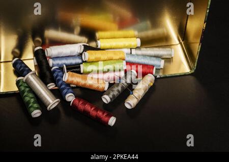 A tin box full of spools of sewing thread upended on a smooth black surface Stock Photo