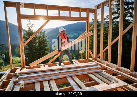 Carpenter constructing wooden-framed house near the woods at sunset. Man holding large joist and dressed in work clothes and helmet. Concept of modern and environmentally-friendly construction. Stock Photo