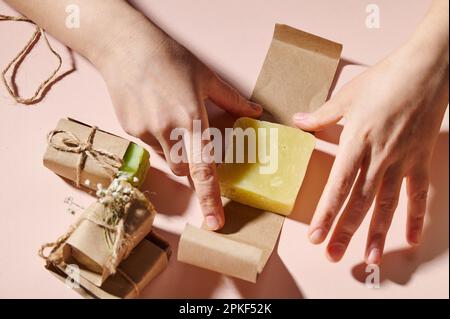 Top view of woman's hands wrapping handmade organic eco soap bar in craft paper, on isolated pink background. Stock Photo