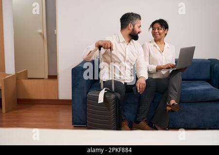 Happy mixed-race tourist couple seated on sofa in hotel room Stock Photo
