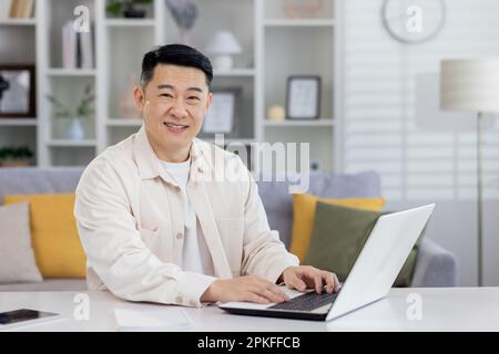 Portrait of successful Asian programmer, man working remotely from home office, businessman smiling and looking at camera using laptop at work sitting in living room at home. Stock Photo