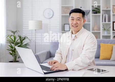 Portrait of successful Asian programmer, man working remotely from home office, businessman smiling and looking at camera using laptop at work sitting in living room at home. Stock Photo