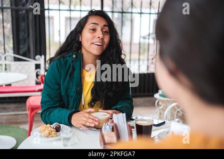 Over the shoulder shot of two latin female friends chatting and drinking latte coffee in a restaurant Stock Photo