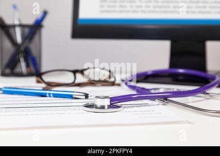 Close-up of a stethoscope over a doctor's white table where are a document, a pen, computer monitor, eyeglasses and pencils pot Stock Photo
