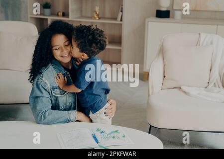 Cute mixed race boy little kid gently hugs his happy mother sitting on floor by table with colored pencils on it, surrounded by light colored furnitur Stock Photo