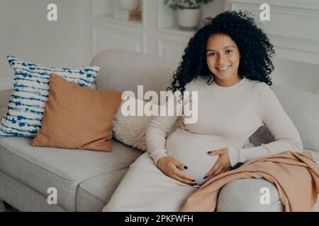 Happy pregnancy time. Cheerful young expectant lovely mixed race young mother with natural looking curly hair relaxing on sofa in living room, touchin Stock Photo