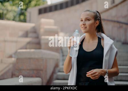 Athlete woman with pony tail feels thirsty after cardio training drinks water from bottle holds refreshig drink stays hydrated poses outdoors dressed Stock Photo