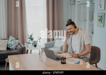 Cheerful bearded young man wearing headphones having online meeting making video call on laptop while working at cozy workplace at home office, sittin Stock Photo