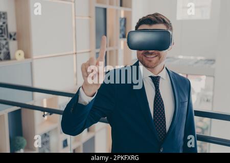 Portrait of young cheerful businessman in formal suit and VR headset goggles explores data visualized in virtual reality and touching it with index fi Stock Photo