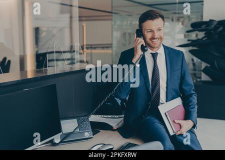 Close-up photo of attractive well-groomed confident businessman in formal wear, speaking on his office desk phone while sitting on top of table, holdi Stock Photo