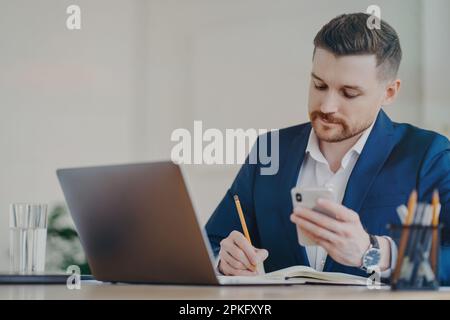Young successful businessman with mobile phone in hand writing down important data into notepad while sitting in front of laptop at office, wearing st Stock Photo
