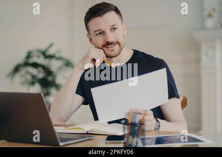 Glad handsome European businessman holds paper document poses in home interior at desktop surrounded by modern technologies checks information has rem Stock Photo