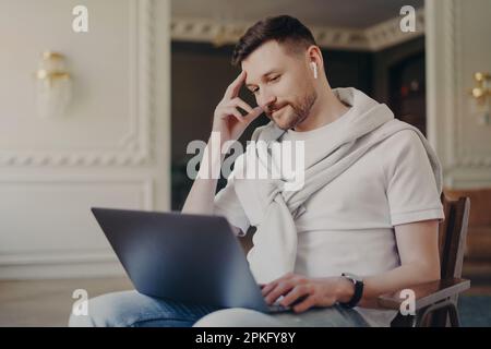 Handsome bearded man in casual clothes concentrated at laptop computer watches webinar communicates in video chat has online conference with client po Stock Photo