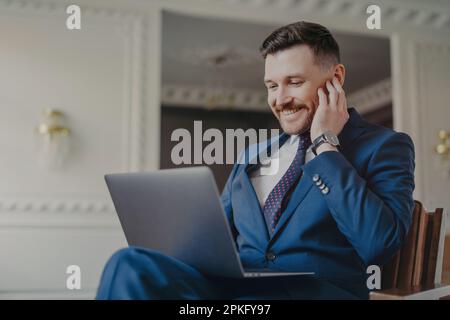 Cheerful corporate employer uses laptop computer reads news online or financial publication connected to 4g internet sits in comfortable armchair dres Stock Photo