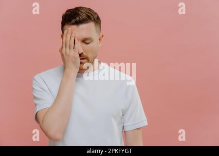 Horizontal shot of serious tired man makes face palm feels bored spending time alone covers eye with hand dressed in casual white t shirt isolated ove Stock Photo