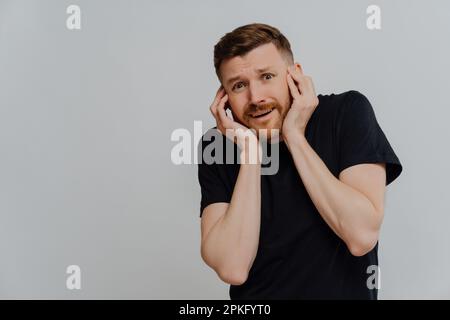 Scared young bearded man in black tshirt holding hands near face and being afraid, looking at something terrible or scaring with horrified facial expr Stock Photo