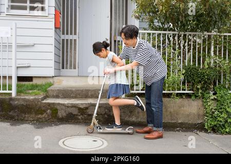 Father teaching his daughter how to ride a kickboard in front of their house Stock Photo