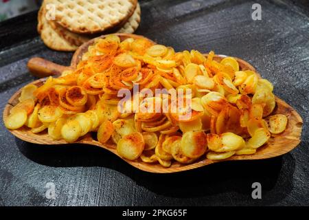 Fried spicy round slices of new potatoes in wooden traditional plate on black oily hob and crunchy flat baked breads at food fair in Bucharest Stock Photo