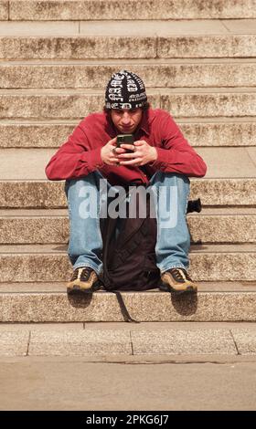 Young man sitting on stone steps in Trafalgar Square, London. UK looking at his phone wearing a wooly black and white hat, red jumper and jeans Stock Photo