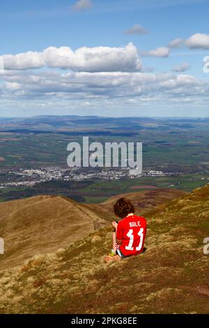 UK Weather: Good Friday bank holiday, April 7th, 2023. Brecon Beacons National Park, South Wales. A boy wearing a Gareth Bale #11 Wales football shirt plays with his smartphone on the summit of Pen y Fan in the Brecon Beacons National Park. The town of Brecon is in the distance. Beautiful sunny weather meant many people made the trip to the Park for today's bank holiday. Stock Photo