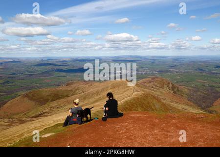 UK Weather: Good Friday bank holiday, April 7th, 2023. Brecon Beacons National Park, South Wales. Hikers looking at their smartphones on the summit of Pen y Fan in the Brecon Beacons National Park. The town of Brecon is in the distance, the ridge in the centre is Cefn Cwm Llwch, the approach route from the north. Beautiful sunny weather meant many people made the trip to the Park for today's bank holiday. Stock Photo