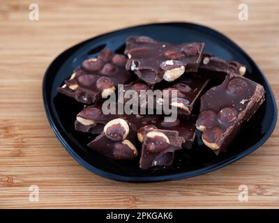Pieces of broken dark chocolate with hazelnut in a black plate. Close up. Stock Photo