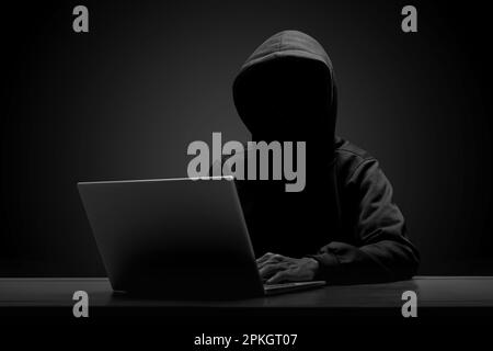 Mysterious man wearing black hoodie using laptop in the dark room. Hacker and cyber security concept. Stock Photo