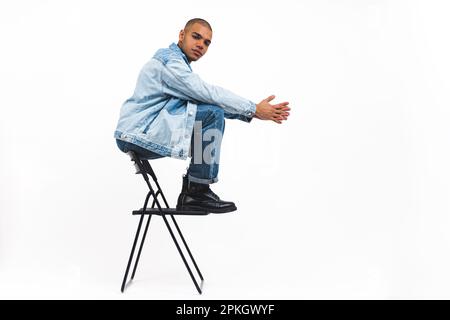 Young adult male model in denim set sitting on a chair in unconventional way. Dangerous poses during fashion photoshoot. High quality photo Stock Photo