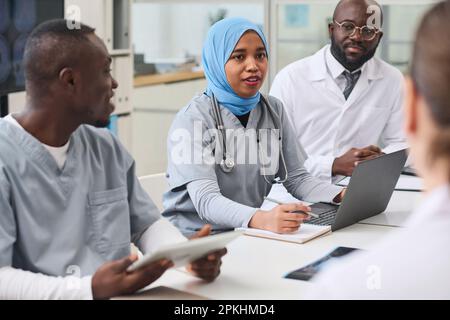 Young female doctor in hijab using laptop and talking to her colleagues during conference in office Stock Photo