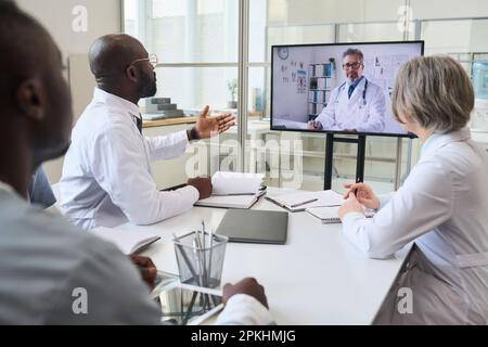 Group of doctors sitting at table and having online meeting with colleague on the monitor, they planning work together Stock Photo