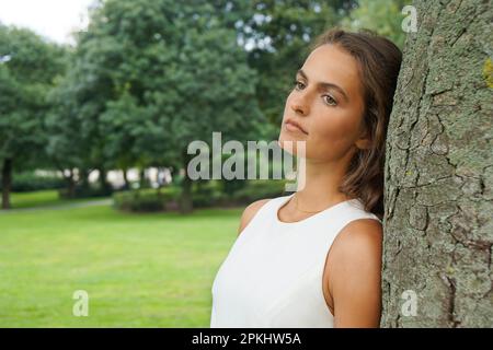 sad young woman leaning against tree gazing absentmindedly Stock Photo