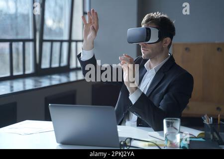 Male office worker in vr headset interacting with digital interface while sitting at desk, touching 3d objects in air, businessman in suit testing vir Stock Photo
