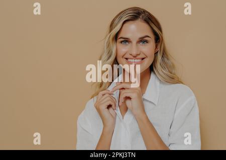 Keep it in secret. Young beautiful positive female with blond dyed hair touching lips with finger and smiling at camera, asking for silence while posi Stock Photo