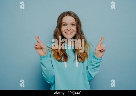 Excited smiling young girl with wavy ombre hair keeping fingers crossed looking at camera wearing casual sweater posing isolated on light blue studio Stock Photo