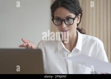Confident woman in glasses sitting at desk, works on computer at home. Concentrated business lady looks at laptop screen gestures to communicate with Stock Photo
