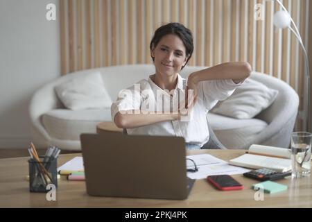 Positive business woman doing hands and fingers stretching exercises while listening to training lectures during webinar online on laptop. Freelancer Stock Photo