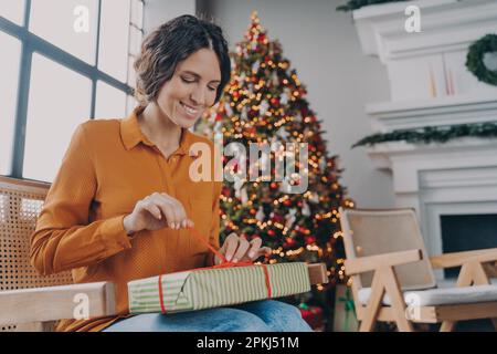 Cheerful happy European woman unpacking Xmas gifts at home, anticipating joyful mood of upcoming winter holidays, smiling Italian female sitting in ro Stock Photo