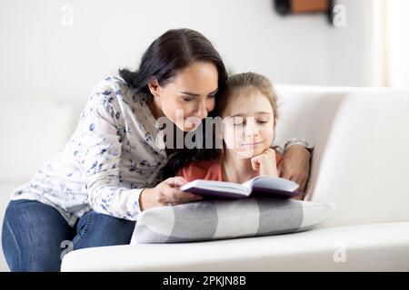 Supportive woman reads a book along with a girl at home on a couch. Stock Photo